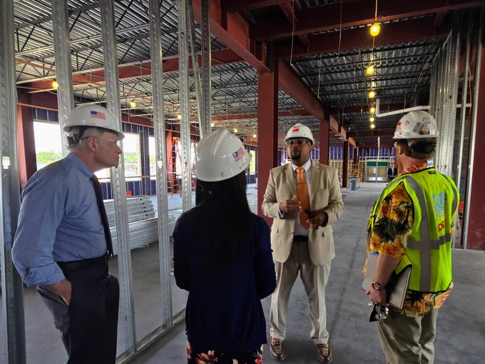 Four people wearing hard hats discuss something in a metal frame construction site, with one person gesturing while speaking.
