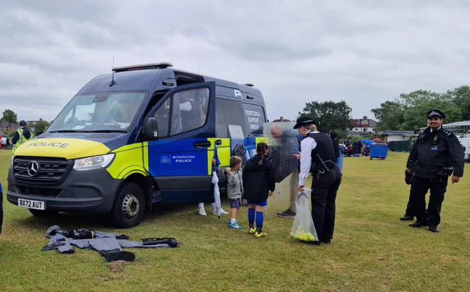 There is a Met Police Territorial Support Group van parked up in between pitches at Wealdstone Football Club, with one of the side doors open, whilst a girls only under 16 football tournament is taking place. The officers have laid out some of their specialist clothing on the ground next to the van and a couple of uniformed officers are showing some young children one of the shields and some of the other kit they have. The uniformed female police officer is also holding a bag of tennis balls.