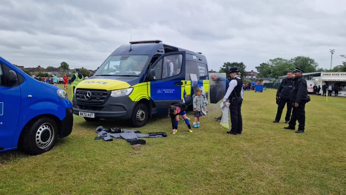There is a Met Police Territorial Support Group van parked up in between pitches at Wealdstone Football Club, with one of the side doors open, whilst a girls only under 16 football tournament is taking place. The officers have laid out some of their specialist clothing on the ground next to the van and a couple of uniformed officers are showing some young children one of the shields and some of the other kit they have. The uniformed female police officer is also holding a bag of tennis balls.