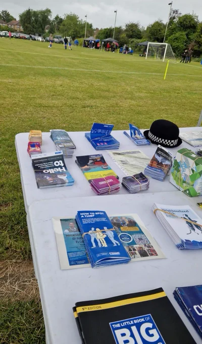 In a space between pitches at Wealdstone Football Club, there is a table with assorted Met Police Crime Prevention materials on, and a female police officers bowler hat also on the table. This is whilst a girls only under 16 football tournament is taking place, and you can see some practising/a match taking place in the background and being watched by a number of spectators.