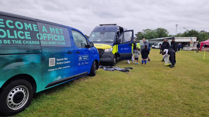 At the front of the photo there is another vehicle advertising Met Police recruitment, which says 'Become a Met Police Officer - Be the change you want to see', and includes a QR  code and that they can be followed via @Met_connected. There is also the Met Police insignia with ' More Trust, Less Crime, High Standards' on the van. At the back of the photo there is a Met Police Territorial Support Group van parked up, with one of the side doors open. The officers have laid out some of their specialist clothing on the ground next to the van and a couple of uniformed officers are showing some young children one of the shields and some of the other kit they have. The uniformed female police officer is also holding a bag of tennis balls. This is between pitches at Wealdstone Football Club whilst a girls only under 16 football tournament is taking place.