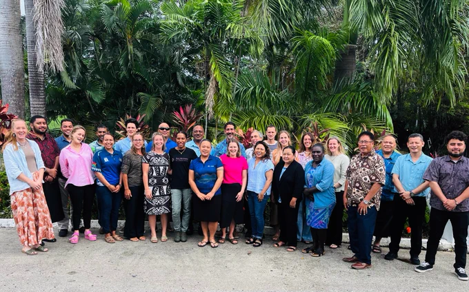 A group of people stand together smiling at the camera. Tropical vegetation in the background