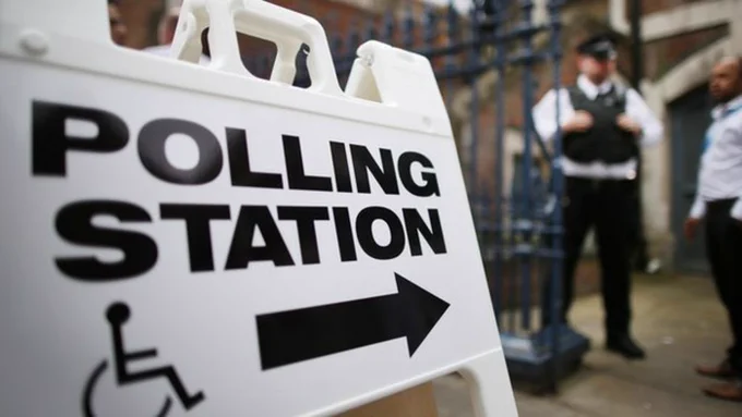 Image shows a white plastic sign with the words "polling station" written upon it with an arrow and the logo of a wheelchair. The arrow is pointing toward the entrance of a polling station where a policeman can just be seen.