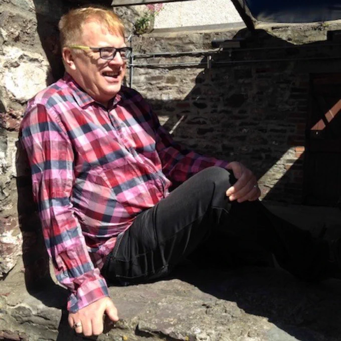 A man sitting on stone steps, wearing a plaid shirt and glasses, smiling in a sunny outdoor setting.