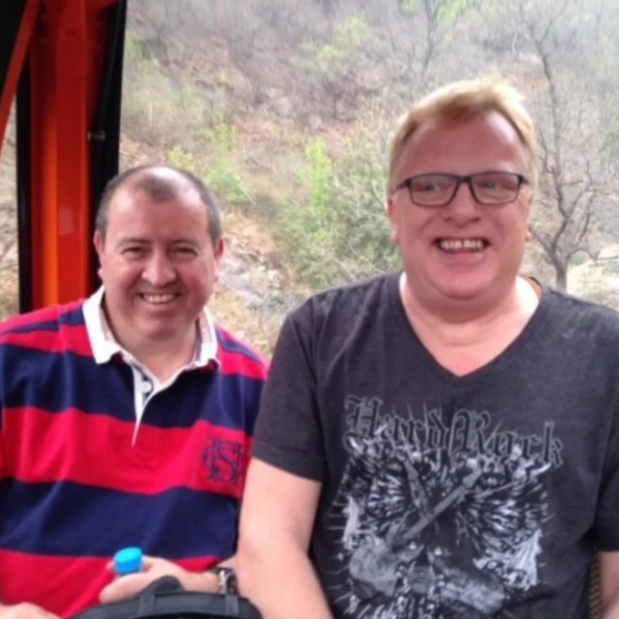 Two men smiling inside a cable car, with natural scenery visible through the window in the background. The person on the left is wearing a striped polo shirt while the person on the right wears a t-shirt with a 'Hard Rock' logo. Both appear happy and are seated during the ride.