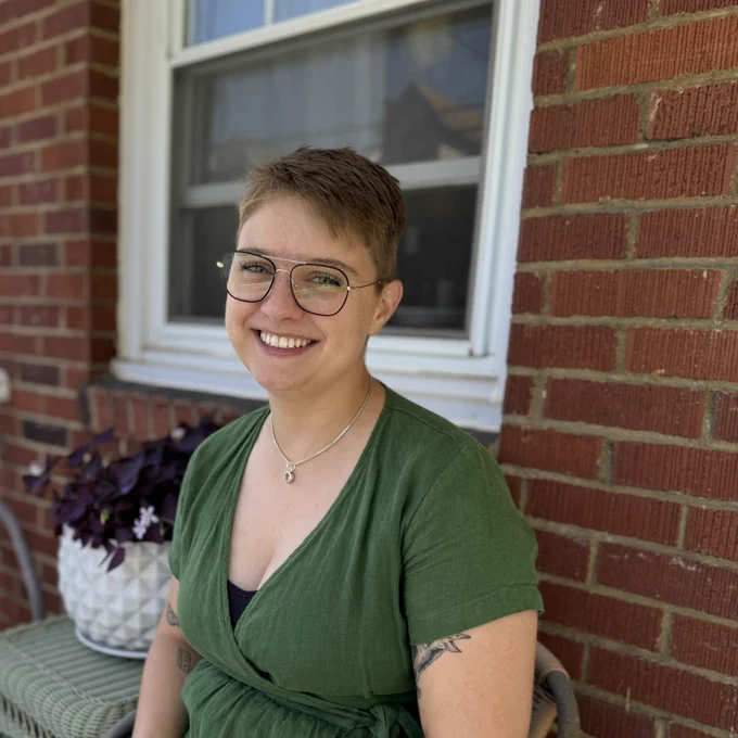 A photo of Alison, a white person with short blonde hair and black wire-framed glasses, wearing a green dress and smiling in front of a brick wall next to a thriving potted plant with purple leaves and tiny flowers. 