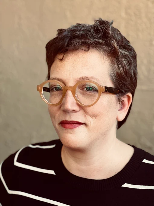 Close-up portrait of a person with short, textured hair and glasses with a thick, camel-colored frame. They are wearing a black and white striped top and have subtle makeup including red lipstick. Their expression is serene, with a slight, thoughtful gaze directed towards the camera.