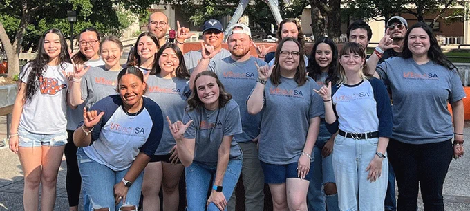 Group of people of various ages wearing "UTSA" t-shirts, posing and smiling in an outdoor setting, some gesturing with their hands in a "birds up" sign.