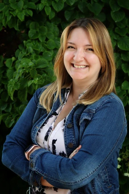 Amber Bedore is smiling at the camera. She is wearing a denim shirt over a floral one and is standing in front of a plant. 