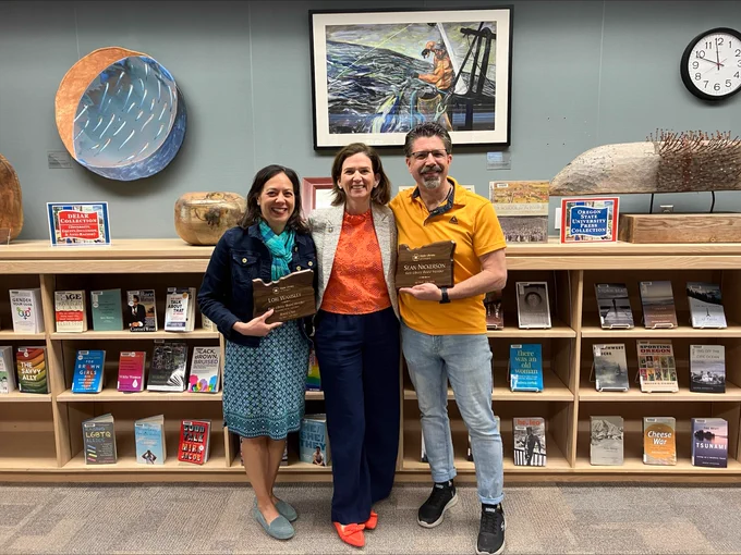 Three people in front of a book shelf, 2 holding awards. 