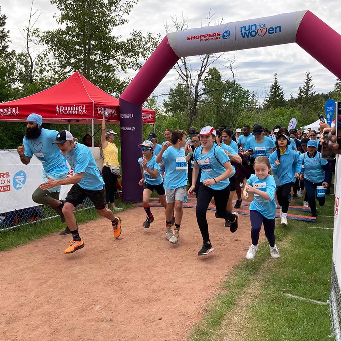 Group of runners in blue shirts crossing the starting line at a charity race event, marked by an overhead arch reading "Shoppers Drug Mart Run for Women."