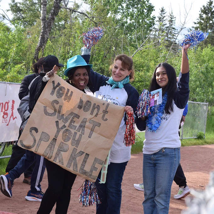 Three people cheerfully participating in an outdoor rally, one holding a sign that says "You don't sweat, you sparkle!" while all holding blue and silver pom-poms, showing enthusiasm and support during the event.