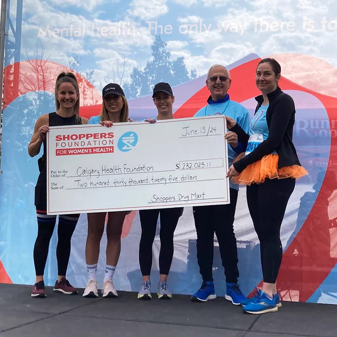 Group of five people holding a large ceremonial cheque at an outdoor event, with a banner in the background reading "Mental health. The only way there is together." The check is for $232,025.11 to the Calgary Health Foundation by Shoppers Drug Mart for Women's Health.