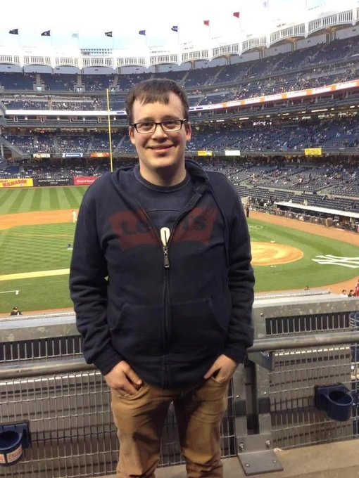 Chris is wearing a Levi's zip up navy blue sweatshirt. He has brown hair and is wearing eyeglasses. He is standing in front of a baseball stadium.