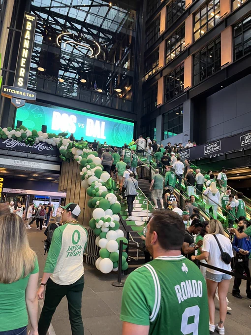 Boston Celtics fans ascend stairs and escalators. On the upper level, a digital screen reads "BOS vs DAL". On the lower level, wayfinding reads "NORTH STATION, COMMUTER RAIL, AMTRAK".