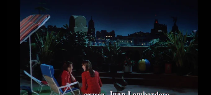 Screenshot de la escena final de Mujeres al Borde de un Ataque de Nervios de Pedro Almodovar.
En la esquina inferior izquierda de la pantalla están sentadas en una reposera Pepa hablando con Marisa en un asiento sin respaldero, ambas vestidas de rojo, en una terraza con una hileras de plantas al fondo en el borde del edificio y de fondo edificios y luces de Madrid. 