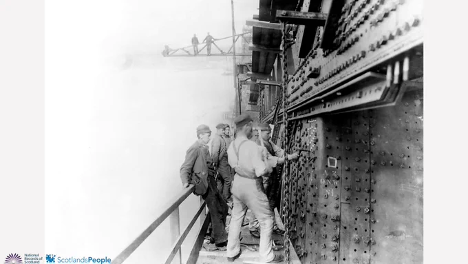 Black and white photograph of riveters in the centre of the photo working on a section of the Forth Bridge, to the right. Mist obscures the background to their left, beyond a guardrail. In the background, two bridge workers look on from a crane.