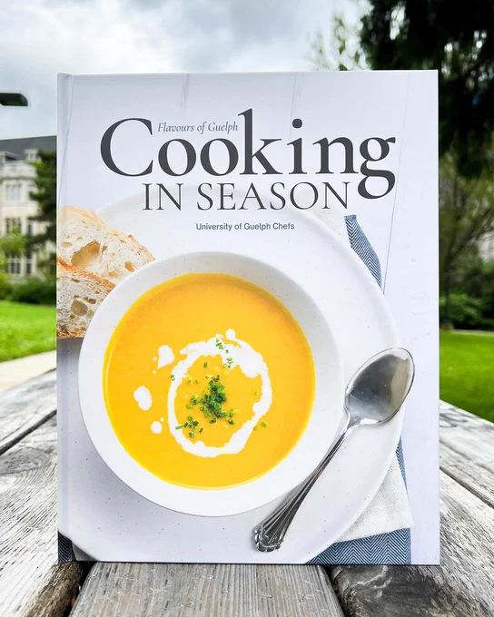 Book set on a table outside, the cover reads "Flavours of Guelph: Cooking in Season, University of Guelph Chefs" and under is a bowl of soup and a spoon.