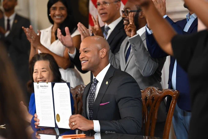 A Black man in a suit seated at a table holds a signed document, smiling, while people behind him stand and applaud.