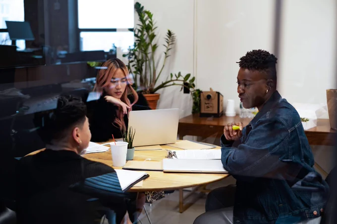 3 professionals having a meeting around a table in an office. There are devices and notebooks open in front of them.
