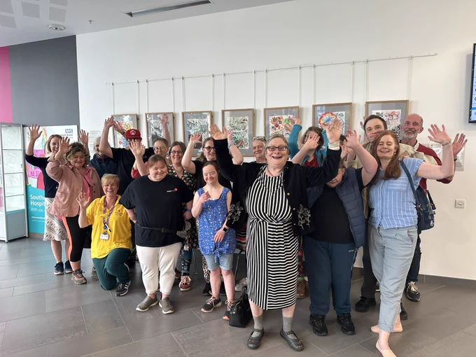 18 people from misfits and creative shift stood together infront of their colourful framed art work all celebrating with their arms in the air.