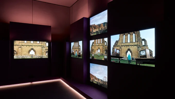 Image: Photograph of a gallery space with purple lighting. There are six screens, one on one wall and five on the other. The images on the screen show buildings. 