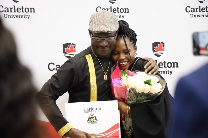 Two individuals embracing joyfully at a Carleton University graduation ceremony. One holds a bouquet of flowers and the other displays a certificate. They are surrounded by banners with the Carleton University logo and Canadian flags.