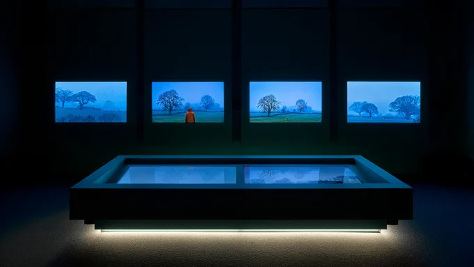 Image: Photograph of a gallery space with four screens showing footage of a hazy blue countryside landscape.