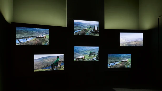 Image: A green-lit gallery with six screens showing footage of people sitting in the countryside on benches.