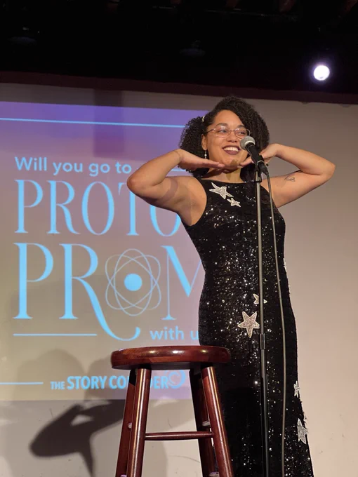 Moiya posing and speaking into a microphone on stage in a black sparkly gown. A screen behind her says Proton Prom