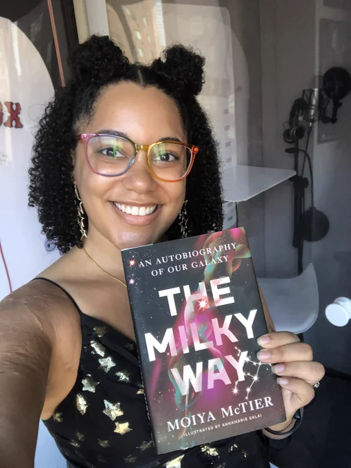 Moiya in rainbow glasses and a star-studded dress smiling and holding a copy of her book, THE MILKY WAY: An Autobiography of Our Galaxy