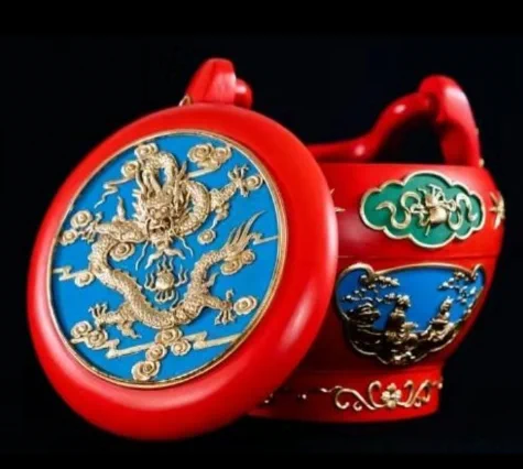 A photo of a traditional craft object in the traditional Chinese art form of Nijincaiqi. It is a red bowl with ornate blue and gold carvings on the lid and sides. On the top is a golden carved dragon. 