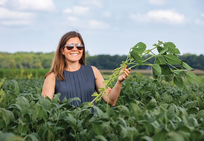 A woman wearing sunglasses surrounded by tall green plants holding a green plant.