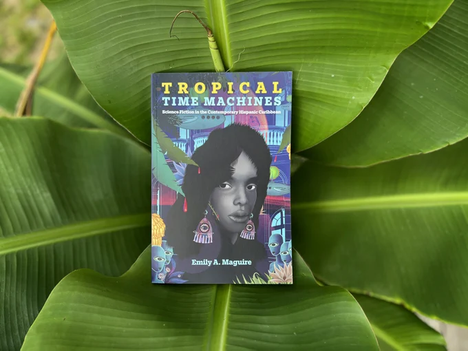 A copy of Tropical Time Machines: Science Fiction in the Contemporary Hispanic Caribbean by Emily A. Maguire sits ensconced in bright green tropical leaves. The cover features a technicolor, sci-fi version of a city as well as a black-and-white depiction of a woman looking to the side with large, colorful dangling earrings.