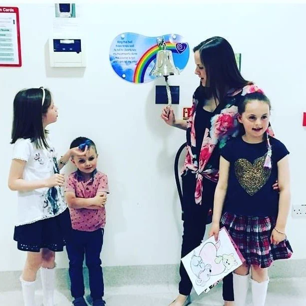 Sarah and her three children in a hallway. Sarah is interacting with one child ringing a bell mounted on the wall, under a rainbow-colored plaque labelled "End of Treatment Bell." 