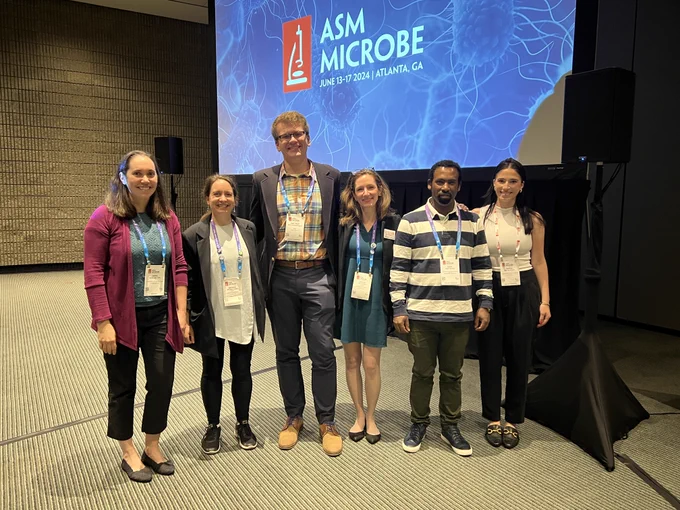 Six speakers standing in a row and smiling in front of a screen displaying the text "ASM Microbe - June 13-17 2024. Atlanta Georgia". From left to right, is Daria Van Tyne, Brooke Talbot, Matthew Anderson, Ashlee Earl, Sisay Worku, and Sophie Nozick.  