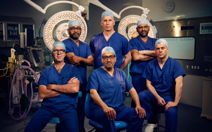 Six surgeons wearing blue scrubs and posing for a group photograph in an operating theatre. 