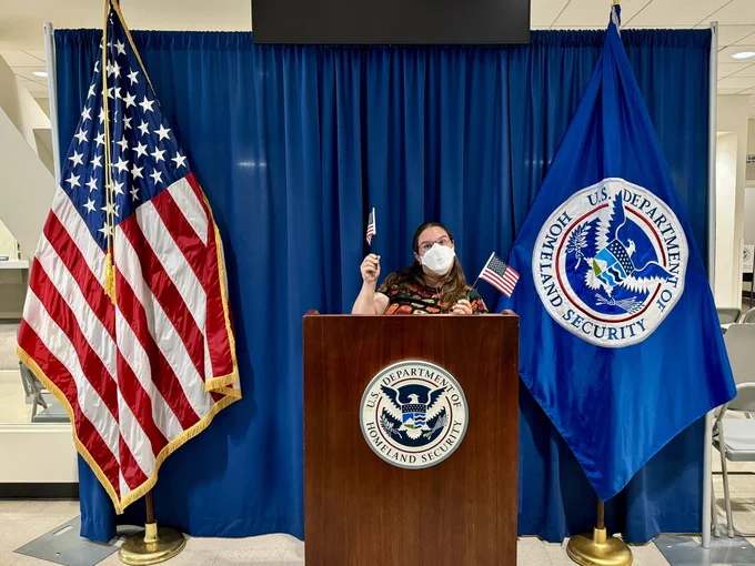 Me standing on a podium near an American flag and the flag for the dept of homeland security, I am waving two tiny American flags whilst wearing a dress with owls on