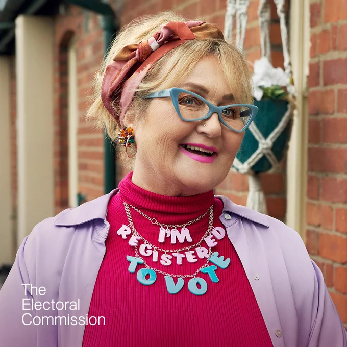 A woman wearing a light purple coat and pink turtleneck with colourful glasses has a pink and blue necklace that reads: "I'm registered to vote."