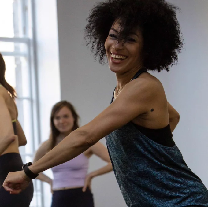 A Black woman with mid-length curly hair and wearing a sleeveless top is caught mid-movement, dancing. She is smiling. A couple more people can be visible in the background. 