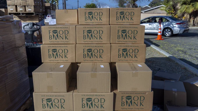 Cardboard boxes labeled "Los Angeles Regional Food Bank" are stacked outdoors for distribution, with a car parked in the background, reflecting an organized food aid event.