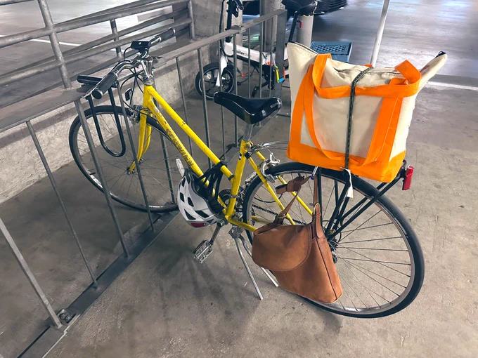 Schwinn caliente yellow 10-speed bike locked up at bike parking rack. A purse is linked on side of rear bike rack and freezer bag bungee’d on top of bike rack.