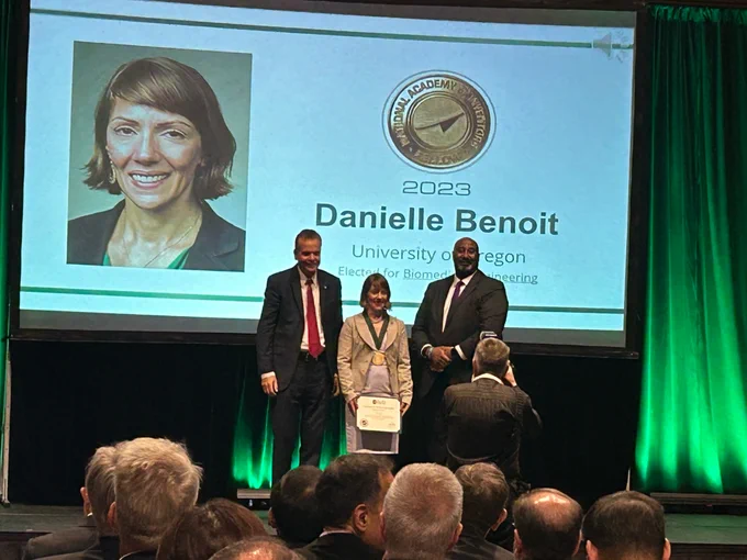 Three individuals on a stage during an award ceremony, with two standing behind a podium. One person is holding an award certificate. A large screen displays a portrait of "Danielle Benoit" and text stating "2023, University of Oregon, Elector for Biomedical Engineering.