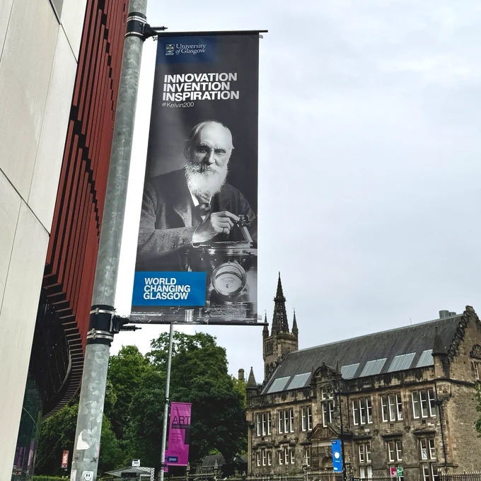 A banner on University Avenue with an archival photo of William Thomson, better known as Lord Kelvin, with text reading: University of Glasgow Innovation Invention Inspiration #Kelvin200 World Changing Glasgow. University buildings and the Tower can be seen in the background with overcast blue skies. 