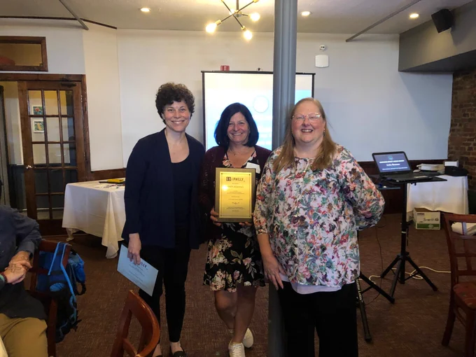 Three people stand indoors, with one holding a plaque. They are smiling and dressed in business casual attire. A presentation setup is visible in the background.