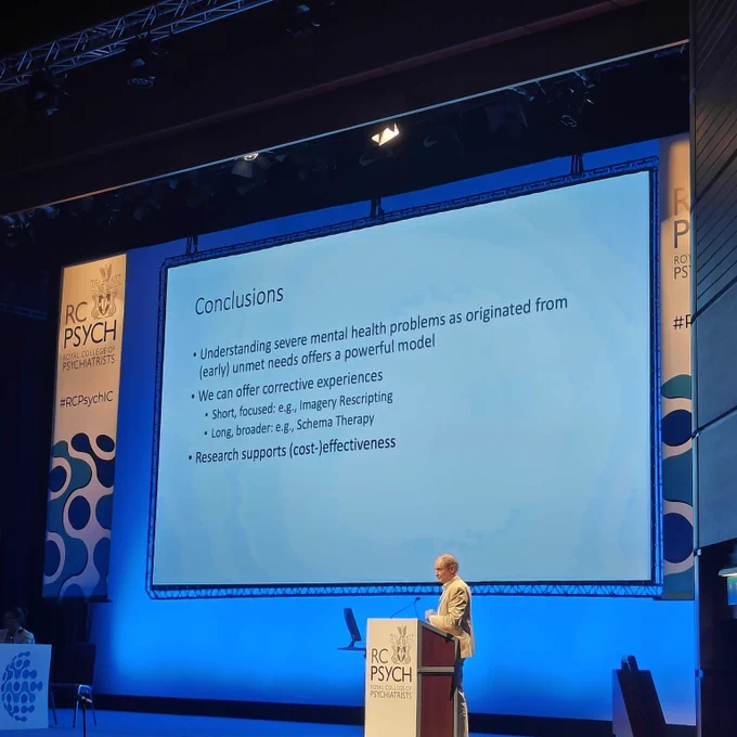 Prof. Emeritus Arnoud Arntz presenting at RC Psych 2024 International Congress, standing at a podium with a large presentation screen behind them displaying a slide titled "Conclusions". The slide outlines points on understanding complex mental health problems, treatment options including Schema Therapy, and research on cost-effectiveness.