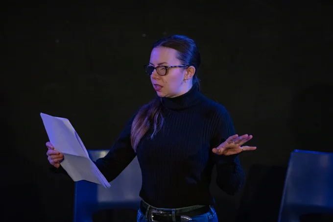 Georgie Wyatt, a white woman with long, dark hair pulled back into a ponytail, standing on a black stage. She is wearing a black turtleneck and reading from a script, one hand out to the side.