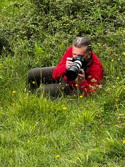 Chris Packham lying down in grass, taking a photo with his camera