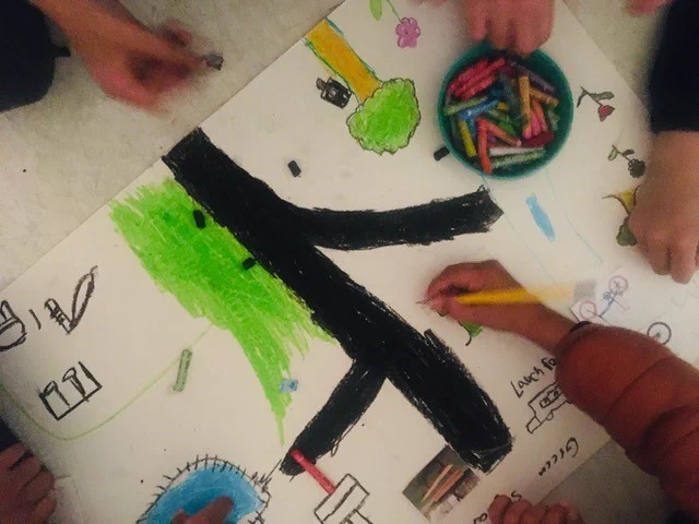 An image of a hand-drawn children's design on paper with flowers, tree and a big path in the middle. Some oil pastels are visible in a tray as well as hands of children holding drawing utensils. 