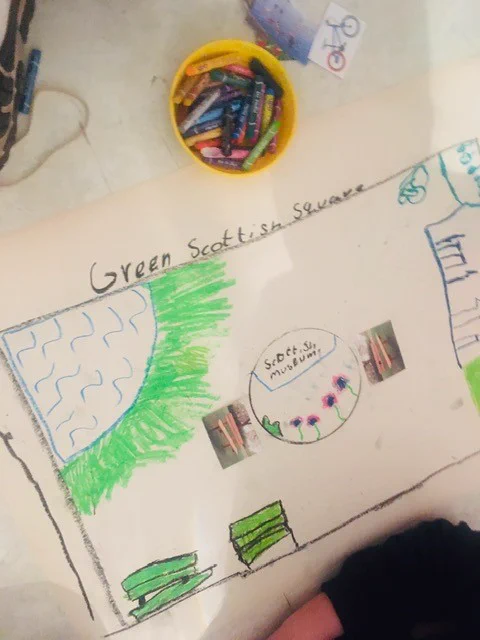An image of a hand-drawn children's design on paper entitled "Green Scottish Square". Some oil pastels are also visible in a tray.